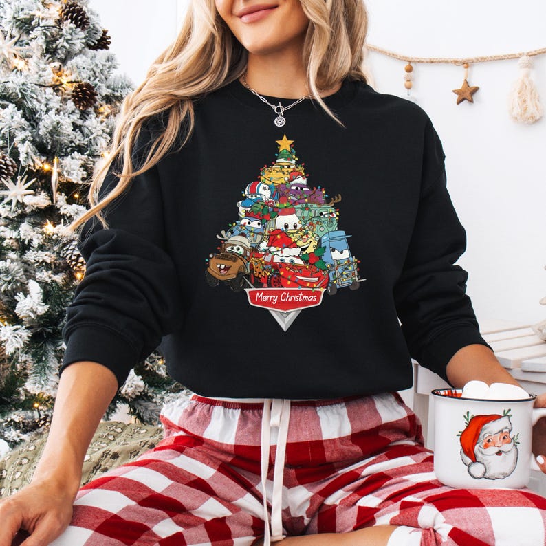 May include: Black sweatshirt featuring a Christmas tree design made of cartoon cars and characters. The tree is topped with a gold star and has the words "Merry Christmas" at the base. The sweatshirt is worn with red and white plaid pajama pants.