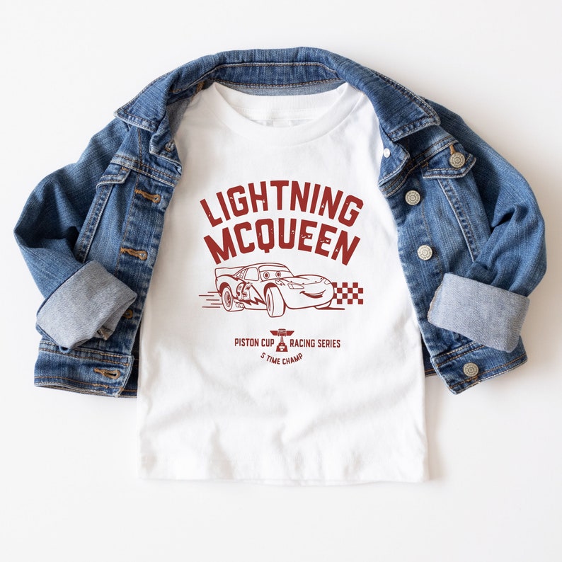 May include: White t-shirt with a red graphic of Lightning McQueen from the Cars movie. The graphic includes the text "Lightning McQueen", "Piston Cup Racing Series", and "5 Time Champ".