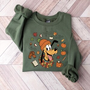 Puede incluir: Sudadera verde con Pluto con gorro naranja rodeado de hojas de oto&ntilde;o, calabazas y bellotas. El dise&ntilde;o incluye una bufanda, una taza de chocolate caliente y un bloc de notas. Las mangas est&aacute;n dobladas.
