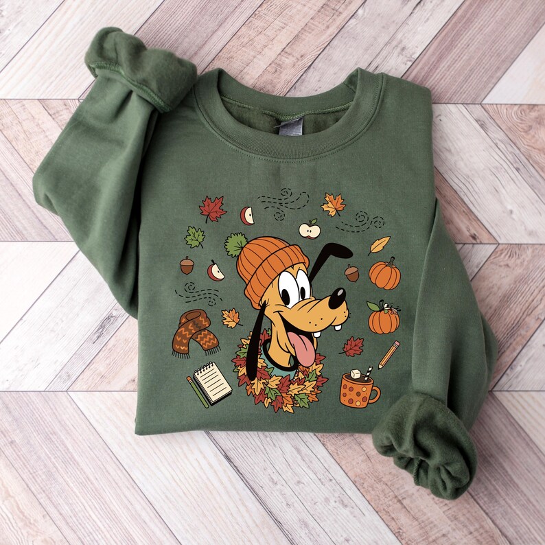 Puede incluir: Sudadera verde con Pluto con gorro naranja rodeado de hojas de oto&ntilde;o, calabazas y bellotas. El dise&ntilde;o incluye una bufanda, una taza de chocolate caliente y un bloc de notas. Las mangas est&aacute;n dobladas.