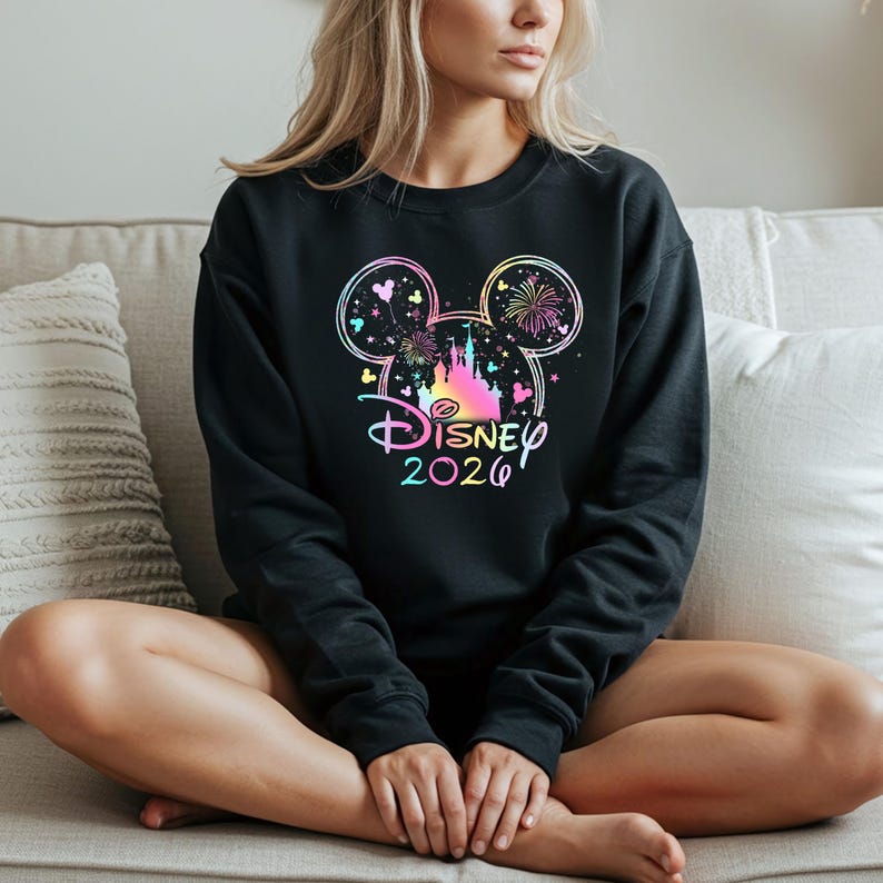 May include: Black crewneck sweatshirt featuring a colorful Disney design. The design includes a castle, fireworks, and the text "Disney 2020" in a rainbow gradient. The sweatshirt is a solid black color.