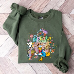 May include: A green sweatshirt with a colorful graphic of Donald Duck in front of the Epcot logo. The graphic also includes a dragon, flowers, and a lollipop.