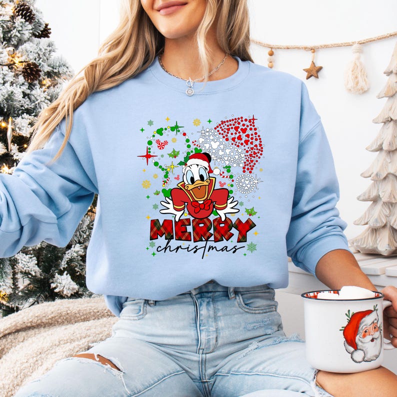 May include: Light blue sweatshirt featuring Donald Duck in a Santa hat and bow tie, with the words "Merry Christmas" in red plaid lettering. The design includes snowflakes and a Santa hat made of hearts.