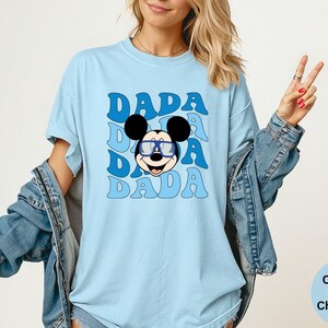 K&ouml;nnte beinhalten: Hellblaues T-Shirt mit dem Wort "DADA" in einem sich wiederholenden Muster und einem Cartoon-Mickey-Mouse-Gesicht mit Brille. Das Shirt ist ein Comfort Colors Chambray.