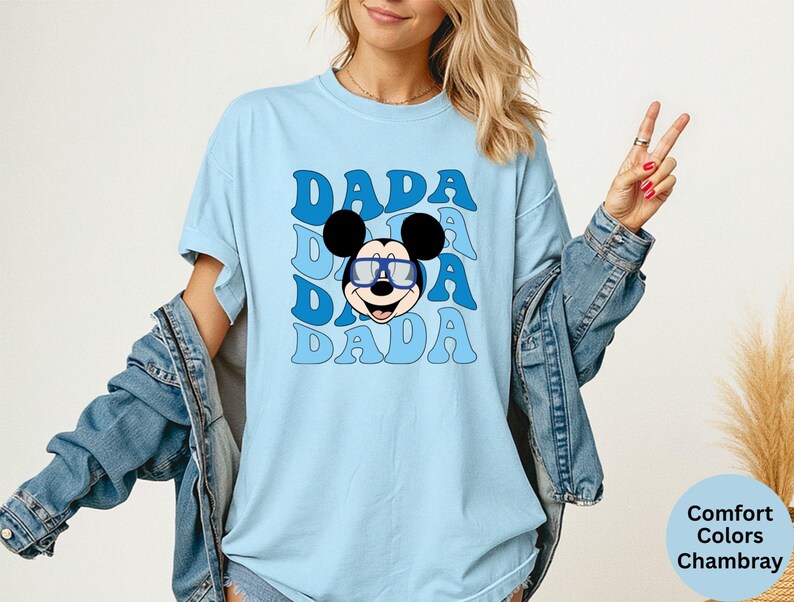 K&ouml;nnte beinhalten: Hellblaues T-Shirt mit dem Wort "DADA" in einem sich wiederholenden Muster und einem Cartoon-Mickey-Mouse-Gesicht mit Brille. Das Shirt ist ein Comfort Colors Chambray.