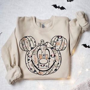 May include: A tan sweatshirt featuring a Halloween-themed Mickey Mouse pumpkin design. The pumpkin is made up of smaller pumpkins and Mickey Mouse faces. The design includes the words "Boo", "Spooky", and "Boo!"