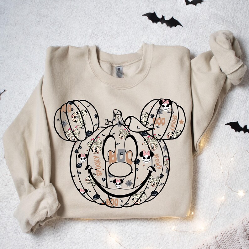 May include: A tan sweatshirt featuring a Halloween-themed Mickey Mouse pumpkin design. The pumpkin is made up of smaller pumpkins and Mickey Mouse faces. The design includes the words "Boo", "Spooky", and "Boo!"