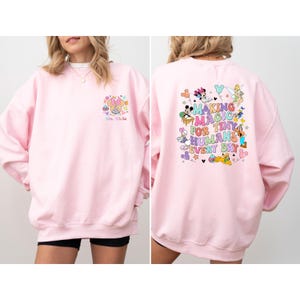 May include: A light pink sweatshirt with a Minnie Mouse graphic on the front and a colorful Disney-themed design on the back. The back design includes the text "MAKING MAGIC FOR TINY HUMANS EVERY DAY".