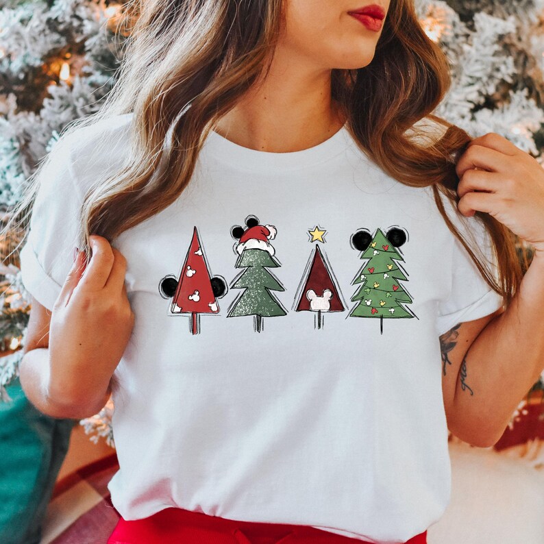 May include: White t-shirt with a graphic of four Christmas trees decorated with Mickey Mouse ears and ornaments. The trees are green, red, and brown.