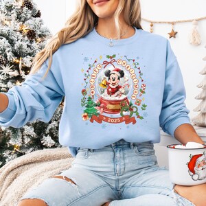 Pode incluir: Camisola azul clara com um design de globo de neve. Mickey Mouse com roupa de Pai Natal segura um sino. O design inclui as palavras "Merry Christmas" e "2025", bengalas doces e &aacute;rvores de Natal.