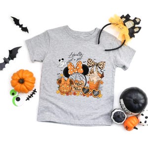 May include: A gray t-shirt with a Halloween-themed graphic featuring Minnie Mouse ears, a pumpkin, a ghost, and a fall-themed drink. The text "Literally just a girl" is above the graphic. Decorative Halloween items surround the shirt.