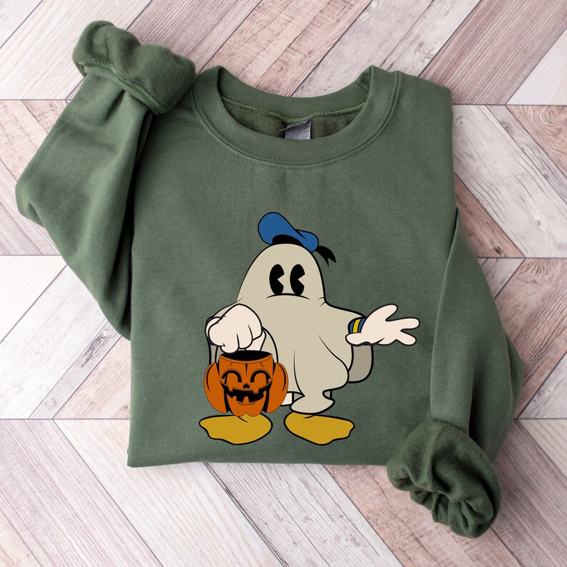 Puede incluir: Una sudadera verde con un fantasma de dibujos animados que lleva un gorro de marinero azul y sostiene una calabaza tallada. El fantasma tiene una expresi&oacute;n de sorpresa y lleva una pulsera amarilla.