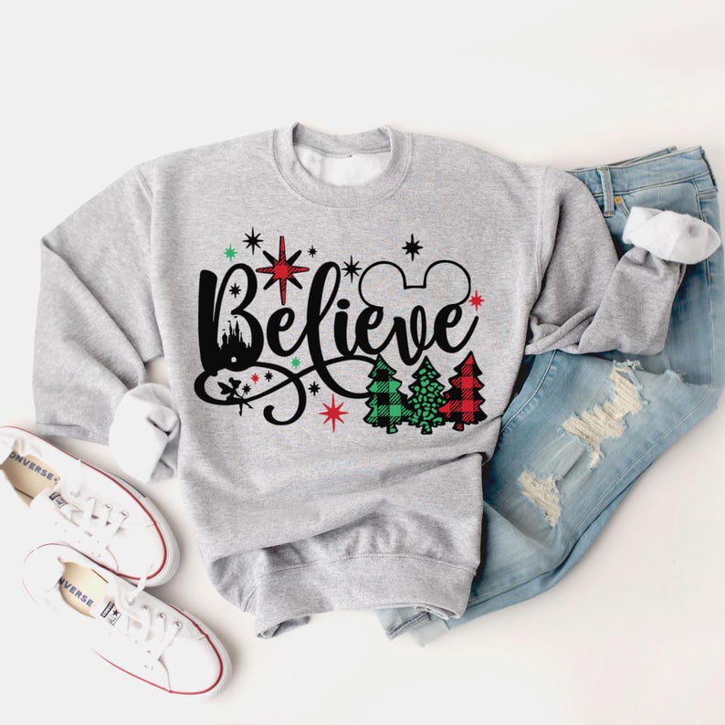 以下が含まれることがあります： 赤い星とミッキーマウスのシルエットがついた、黒い文字で「Believe」と書かれたグレーのスウェットシャツ。スウェットシャツには、赤と緑のチェック柄のクリスマスツリーのデザインも施されています。