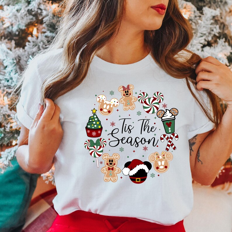 Peut inclure: T-shirt blanc avec un motif de couronne de No&euml;l. La couronne comprend des personnages en pain d'&eacute;pices, des cannes en sucre et un cupcake en forme de sapin de No&euml;l. Le texte "Tis The Season" est au centre.