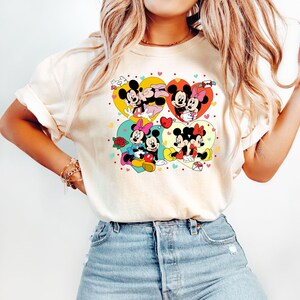 May include: A white t-shirt with a graphic print of Mickey Mouse and Minnie Mouse in four different heart-shaped designs. The hearts are in shades of yellow, red, blue, and green. The graphic is surrounded by small hearts in various colors.
