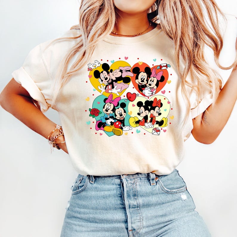 May include: A white t-shirt with a graphic print of Mickey Mouse and Minnie Mouse in four different heart-shaped designs. The hearts are in shades of yellow, red, blue, and green. The graphic is surrounded by small hearts in various colors.