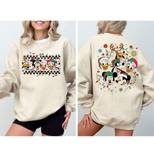 Puede incluir: Una sudadera beige con un dise&ntilde;o navide&ntilde;o con Mickey Mouse, Minnie Mouse, Donald Duck, Goofy y Pluto. El dise&ntilde;o en la parte delantera muestra a los personajes en fila con luces de Navidad y un patr&oacute;n a cuadros. El dise&ntilde;o en la parte posterior muestra a los personajes reunidos alrededor de un &aacute;rbol de Navidad con luces y copos de nieve.
