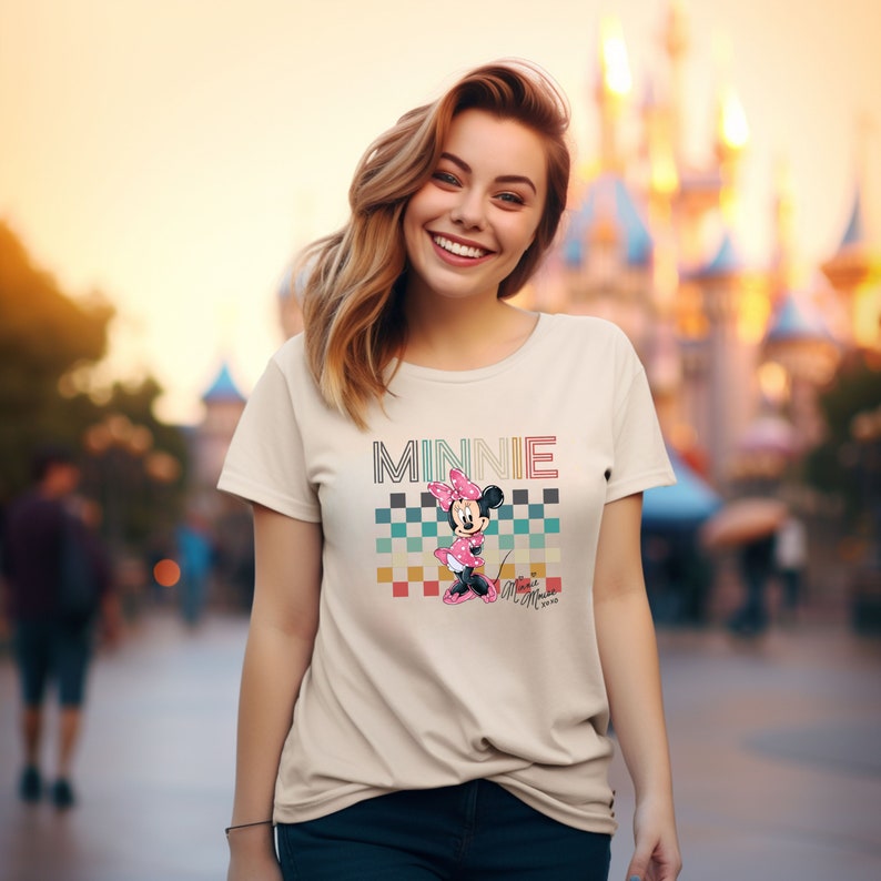 May include: A beige t-shirt with a retro-style graphic of Minnie Mouse. The graphic features Minnie Mouse in a pink dress and bow, standing on a checkered background with the word "MINNIE" in colorful letters.
