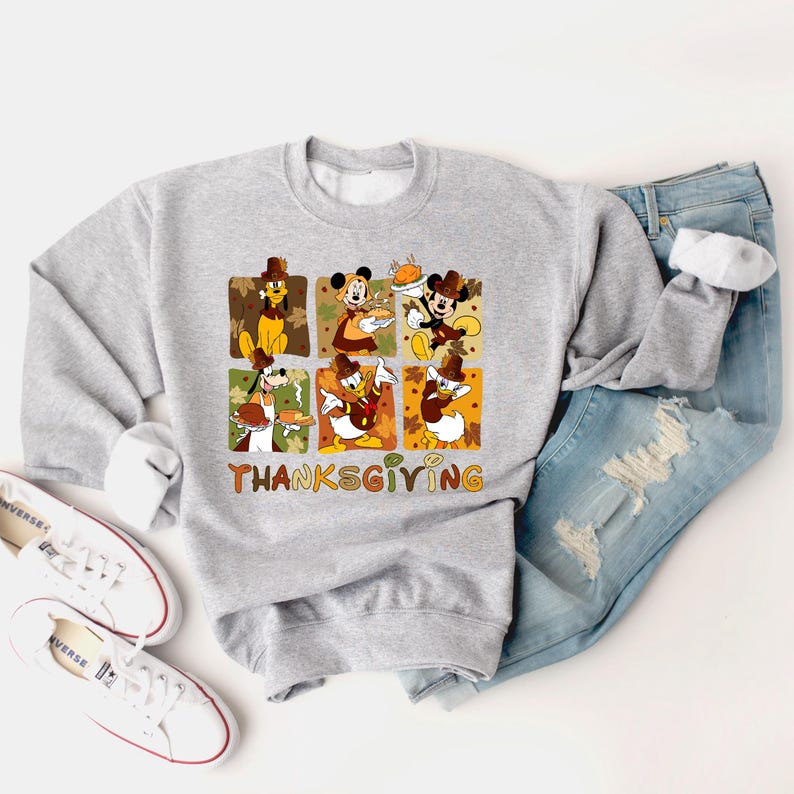 May include: A light gray sweatshirt featuring a Thanksgiving-themed graphic with Disney characters. The graphic includes images of Mickey Mouse, Minnie Mouse, Donald Duck, and Goofy, with the word "THANKSGIVING" in orange.