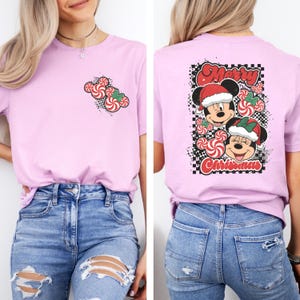 May include: A light pink t-shirt featuring a Christmas design. The front of the shirt has a candy cane graphic. The back of the shirt has a graphic of Mickey and Minnie Mouse wearing Santa hats, with the words "Merry Christmas".