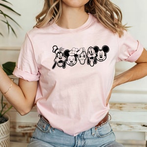 K&ouml;nnte beinhalten: Ein pinkfarbenes T-Shirt mit einer schwarzen Grafik von Mickey Mouse, Minnie Mouse, Donald Duck und Goofy. Die Figuren sind in einer Reihe angeordnet und l&auml;cheln.