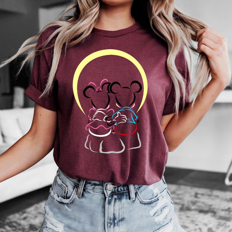 Puede incluir: Camiseta burdeos de manga corta con un gr&aacute;fico de dos personajes de dibujos animados abraz&aacute;ndose, sobre un c&iacute;rculo amarillo. Los personajes est&aacute;n delineados en negro con acentos rosas y azules. La camiseta tiene mangas enrolladas.
