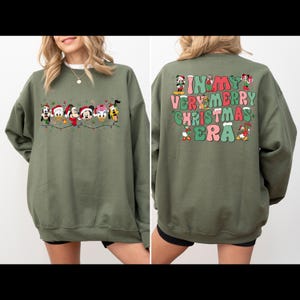 May include: A green sweatshirt with a Christmas-themed design featuring Mickey Mouse and friends on the front and the text "In My Very Merry Christmas Era" on the back.