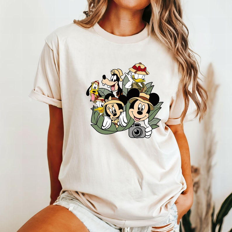 May include: A white t-shirt with a graphic print of Mickey Mouse, Minnie Mouse, Goofy, Donald Duck, and Pluto dressed in safari gear. The characters are surrounded by green leaves and are holding a camera.