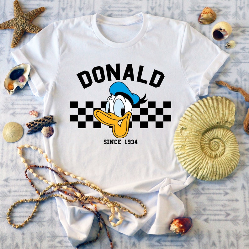 May include: White t-shirt featuring a graphic of Donald Duck with the name "DONALD" above and "SINCE 1934" below. The design includes a checkered pattern and Donald Duck's face with a blue hat and yellow beak.