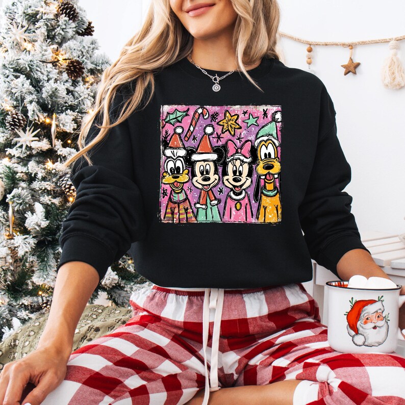 Op de afbeelding: Zwarte sweatshirt met een kleurrijke afbeelding van Disney-personages, waaronder Mickey en Minnie Mouse, Goofy en Donald Duck, die kerstmutsen dragen. Het ontwerp bevat zuurstokken en sterren. De persoon draagt een rood-witte geruite pyjamabroek.