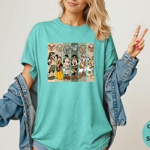 May include: A seafoam green t-shirt featuring a graphic of cartoon characters in safari outfits. The graphic includes Mickey Mouse, Donald Duck, Goofy, and Pluto. The shirt is a Comfort Colors brand.
