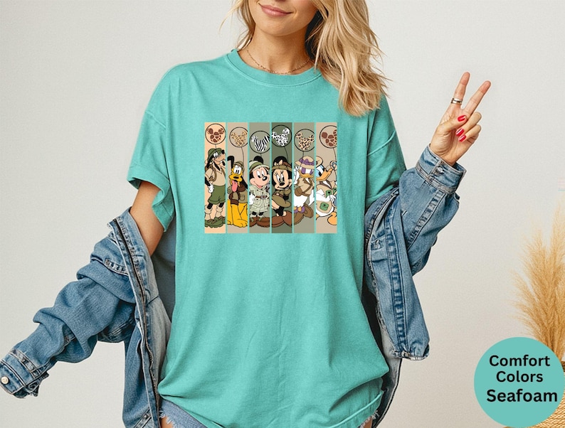 May include: A seafoam green t-shirt featuring a graphic of cartoon characters in safari outfits. The graphic includes Mickey Mouse, Donald Duck, Goofy, and Pluto. The shirt is a Comfort Colors brand.