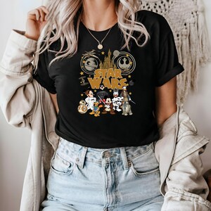May include: Black t-shirt featuring a gold "Star Wars" logo with iconic imagery. The design includes cartoon characters like Mickey Mouse, Donald Duck, and Goofy dressed as Star Wars characters. The shirt has a classic crew neck.