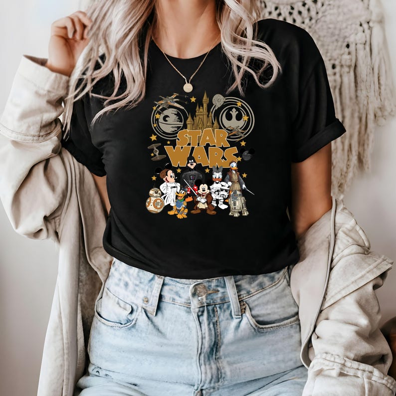 May include: Black t-shirt featuring a gold "Star Wars" logo with iconic imagery. The design includes cartoon characters like Mickey Mouse, Donald Duck, and Goofy dressed as Star Wars characters. The shirt has a classic crew neck.