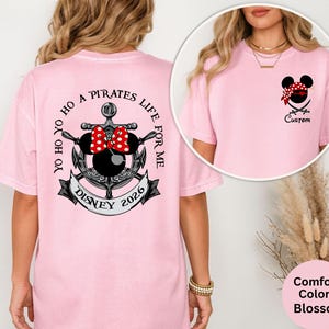 May include: A light pink t-shirt featuring a Minnie Mouse pirate design with a red and white polka dot bow, a bandana, and crossed swords. The back of the shirt has an anchor design with the text "Yo Ho Ho A Pirates Life For Me" and "Disney 2026".