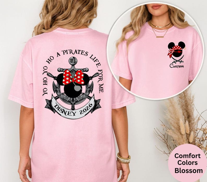 May include: A light pink t-shirt featuring a Minnie Mouse pirate design with a red and white polka dot bow, a bandana, and crossed swords. The back of the shirt has an anchor design with the text "Yo Ho Ho A Pirates Life For Me" and "Disney 2026".