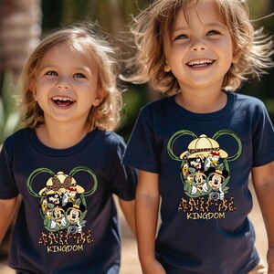 May include: Two children wearing navy blue t-shirts with a Mickey Mouse design. The shirts feature Mickey Mouse and his friends dressed in safari gear with the text "Adventure Kingdom".