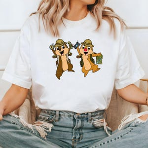 May include: White t-shirt featuring a graphic of two cartoon chipmunks wearing hats and backpacks, holding a compass and a camera. The chipmunks are smiling and appear to be on an adventure.