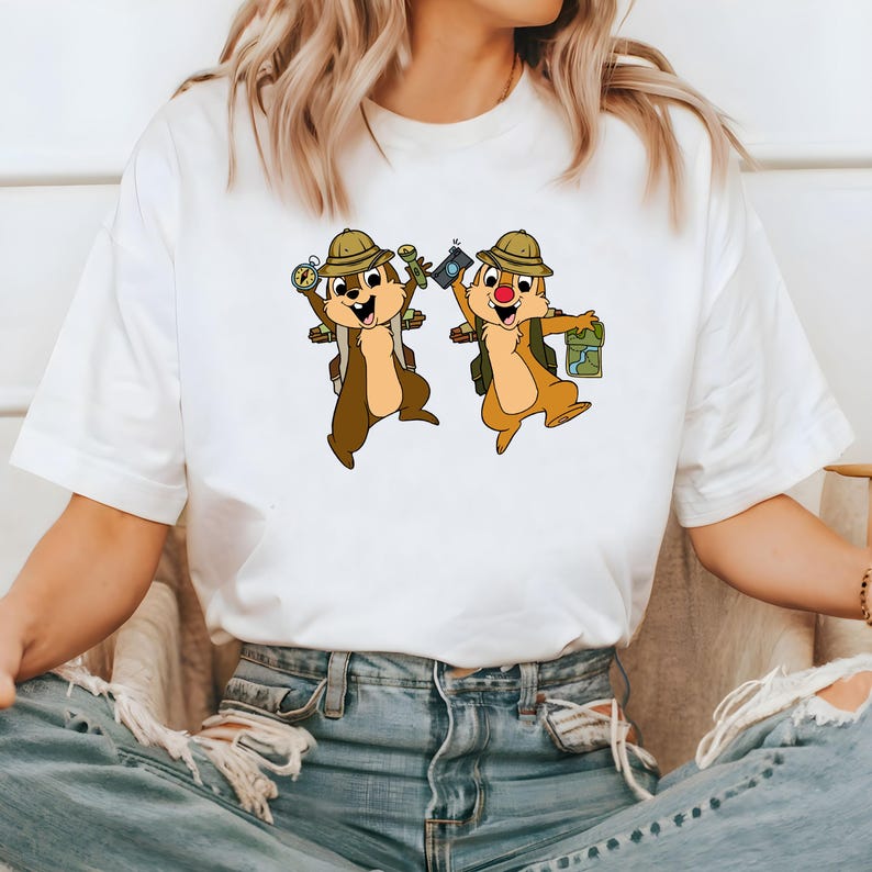 May include: White t-shirt featuring a graphic of two cartoon chipmunks wearing hats and backpacks, holding a compass and a camera. The chipmunks are smiling and appear to be on an adventure.