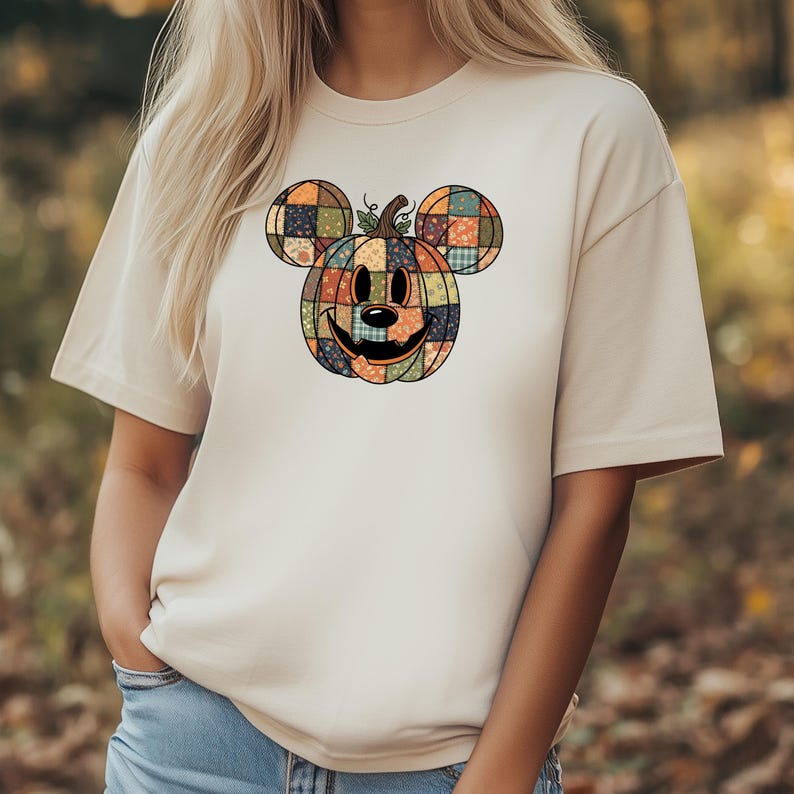 May include: Beige t-shirt featuring a patchwork pumpkin design with Mickey Mouse ears. The pumpkin is composed of various colorful squares and has a smiling face. The shirt is a casual, short-sleeved style.