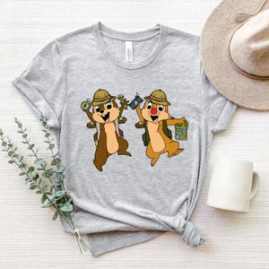 May include: Grey t-shirt featuring a graphic of two cartoon chipmunks wearing explorer hats and carrying backpacks. The chipmunks are holding a compass, a camera, and a map.