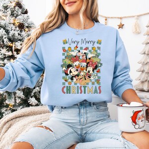 May include: Light blue sweatshirt featuring a colorful graphic of Disney characters, including Mickey and Minnie Mouse, Donald Duck, and Goofy, with the text "Very Merry Christmas." The design includes Christmas trees and gift boxes. A festive holiday-themed apparel item.
