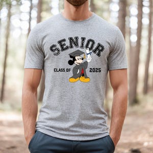 Op de afbeelding: Grijs T-shirt met een zwart-witte afbeelding van Mickey Mouse die een afstudeerpet en toga draagt. De tekst "SENIOR" staat in een zwarte boog boven Mickey Mouse. De tekst "CLASS OF 2025" staat onder Mickey Mouse.