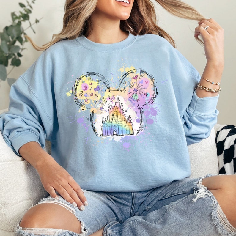 Puede incluir: Una sudadera azul claro con un dise&ntilde;o de cabeza de Mickey Mouse de colores arco&iacute;ris que presenta un boceto del castillo de Cenicienta. El dise&ntilde;o est&aacute; rodeado de salpicaduras de pintura de colores.