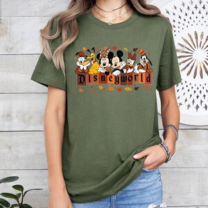 May include: Olive green t-shirt featuring Disney characters Donald Duck, Goofy, Minnie Mouse, Mickey Mouse, Daisy Duck, and Pluto. The word "DISNEYWORLD" is printed in brown letters with autumn leaves.