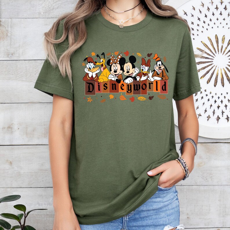 May include: Olive green t-shirt featuring Disney characters Donald Duck, Goofy, Minnie Mouse, Mickey Mouse, Daisy Duck, and Pluto. The word "DISNEYWORLD" is printed in brown letters with autumn leaves.