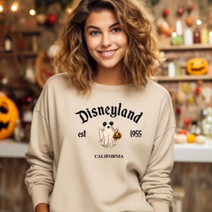 Puede incluir: Una sudadera de color beige con un gr&aacute;fico negro de un fantasma sosteniendo una calabaza. El gr&aacute;fico tambi&eacute;n incluye el texto "Disneyland est 1955 California".