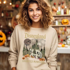 May include: A beige sweatshirt with a vintage-style graphic of Mickey Mouse and a ghost in front of the Haunted Mansion at Disneyland. The text "Disneyland" and "The Haunted Mansion" are printed on the sweatshirt.