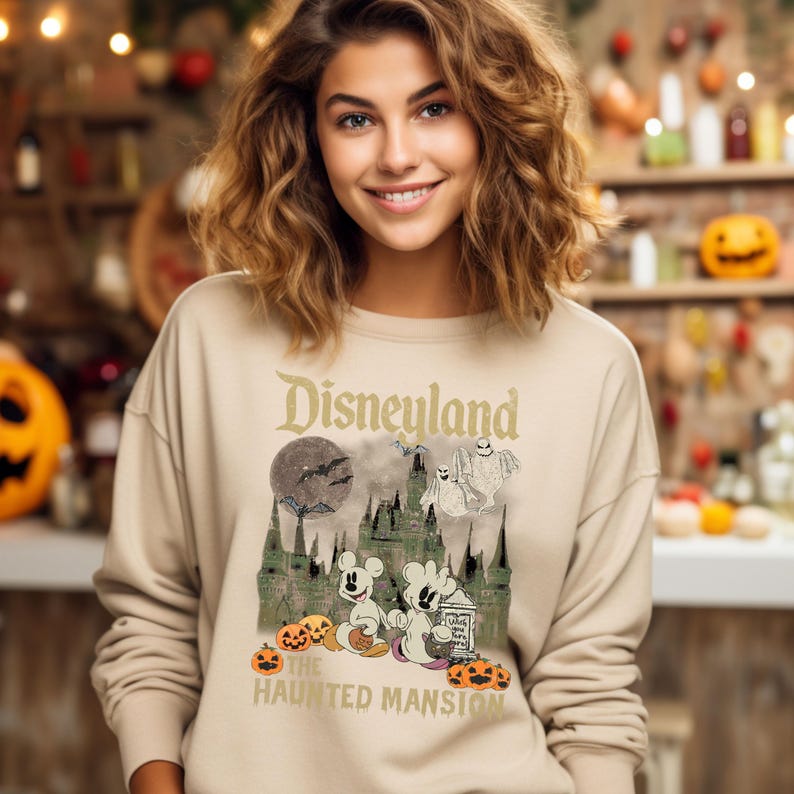 May include: A beige sweatshirt with a vintage-style graphic of Mickey Mouse and a ghost in front of the Haunted Mansion at Disneyland. The text "Disneyland" and "The Haunted Mansion" are printed on the sweatshirt.