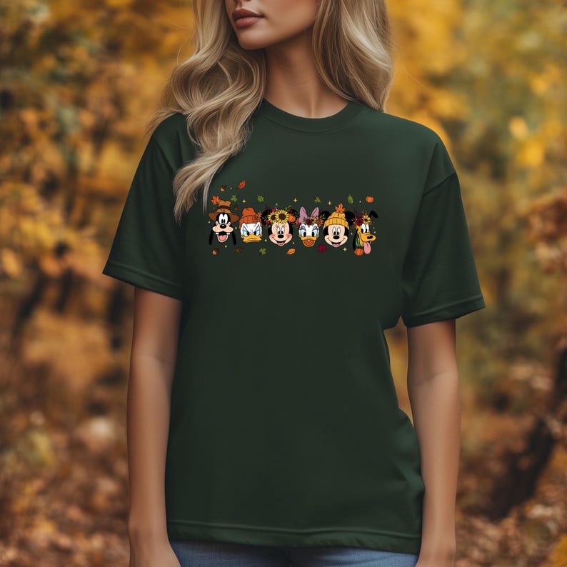 May include: Dark green t-shirt featuring a line of cartoon characters, including Mickey Mouse, Goofy, Donald Duck, Daisy Duck, and Minnie Mouse, each adorned with fall-themed accessories. The design includes autumn leaves.